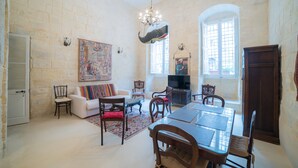 Un salón comedor amplio con techo alto, paredes de piedra, una araña y una televisión. Incluye zona de comedor con muebles de madera.