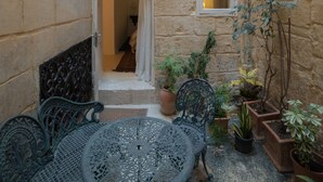 Un patio estrecho con paredes de piedra, una mesa y sillas de metal, y plantas en macetas.