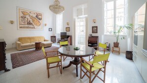 Un salón luminoso con una mesa redonda de vidrio rodeada de sillas amarillas, un sofá color beige y un cuadro grande en la pared.