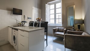 A compact living space with a kitchenette, a washing machine, a dining area with chairs, a sofa, and a large window providing natural light. - JM - Casa Rooms (Valletta)