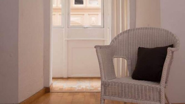 Ein Zimmer mit einem weißen Korbstuhl, einer weißen Tür und Blick auf ein Gebäude durch die Tür.