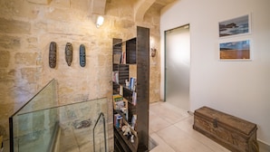 A stone-walled hallway with a glass railing, a wooden chest, and a bookshelf filled with books and decorative items. - TA - Casa Rooms (Valletta)