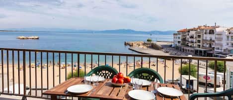 Une table avec des assiettes, des couverts et un saladier de fruits, placée sur un balcon donnant sur une plage, avec des personnes et des bâtiments en arrière-plan.