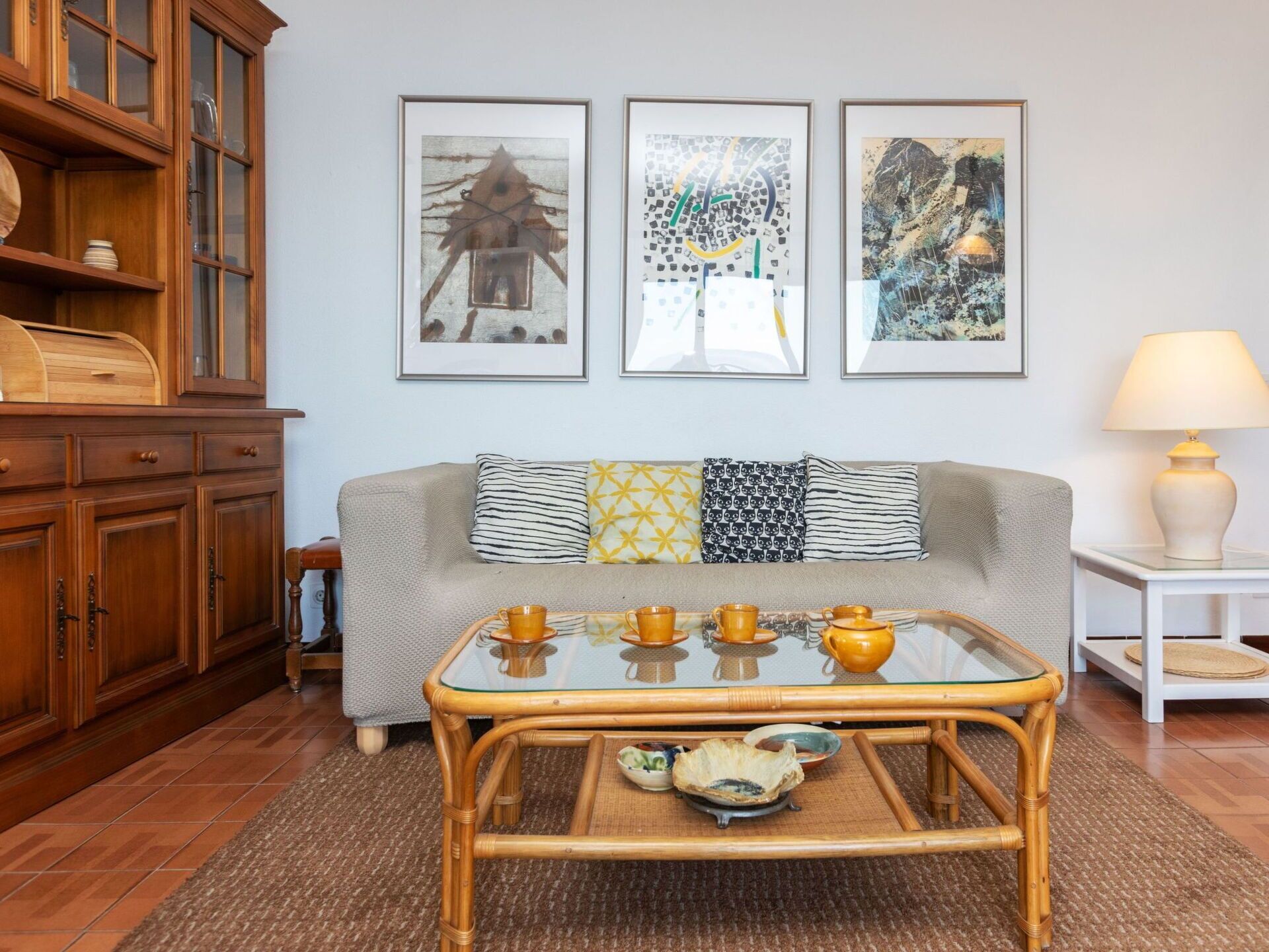 A living room with a sofa, a glass-top coffee table with tea cups, a wooden cabinet, and three framed artworks on the wall.