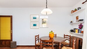 Een eethoek met een ronde houten tafel, zes stoelen en een fruitschaal. Er hangen afbeeldingen in de muur, een hanglamp en een plank met diverse spullen.
