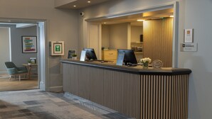 A reception desk with a computer, a small plant, and a sign about laundry pickup. There is a chair and a framed picture on the wall. - Cambridge Bar Hill Hotel, BW Signature Collection (Cambridge)