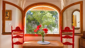 A dining area with a wooden table and two red chairs, a vase with red flowers, and a window with a view of trees. - Cosy apartment for 3 people with pool, WIFI, TV and terrace (Denia)