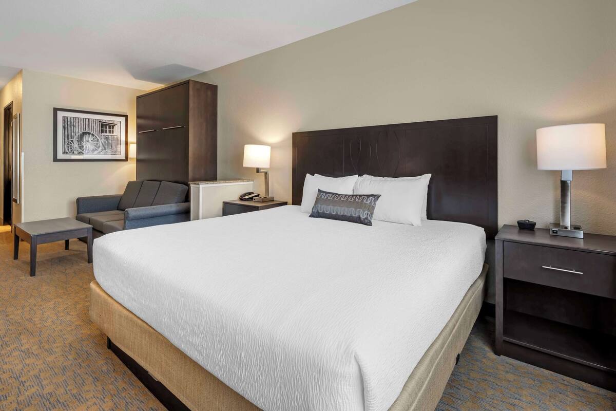 A hotel room with a large bed, a desk, a chair, a wardrobe, and a nightstand with a lamp.