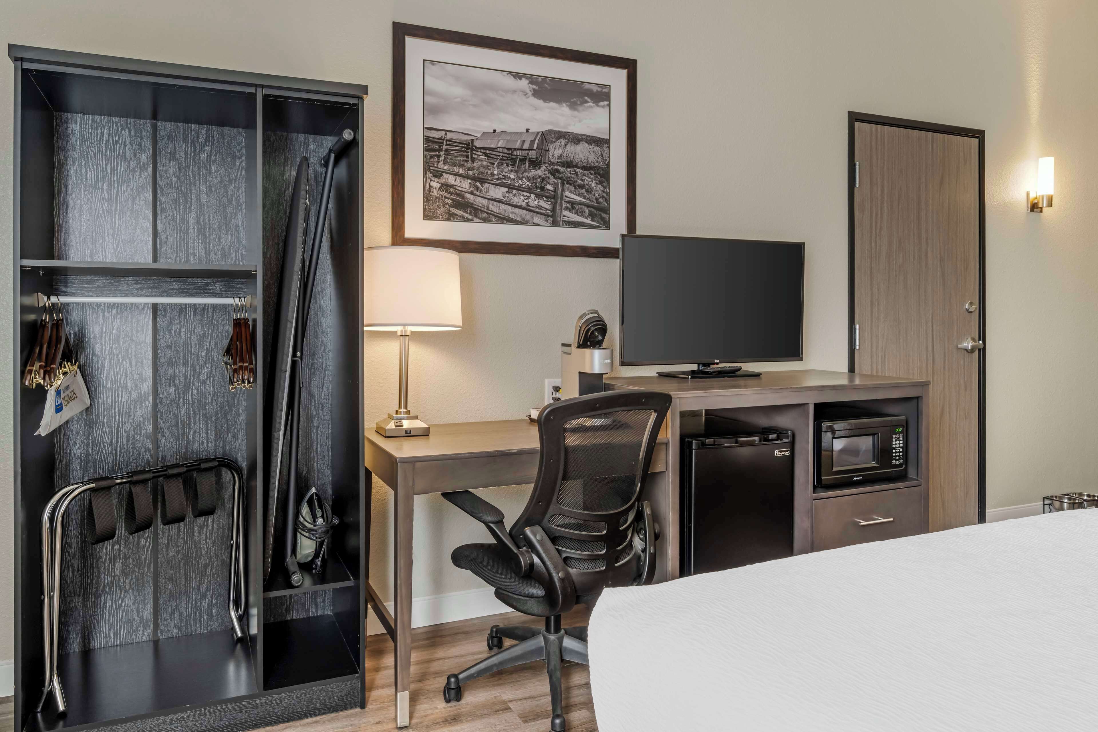 A hotel room with a bed, a desk with a chair, a television, a microwave, and a wardrobe with hanging clothes.