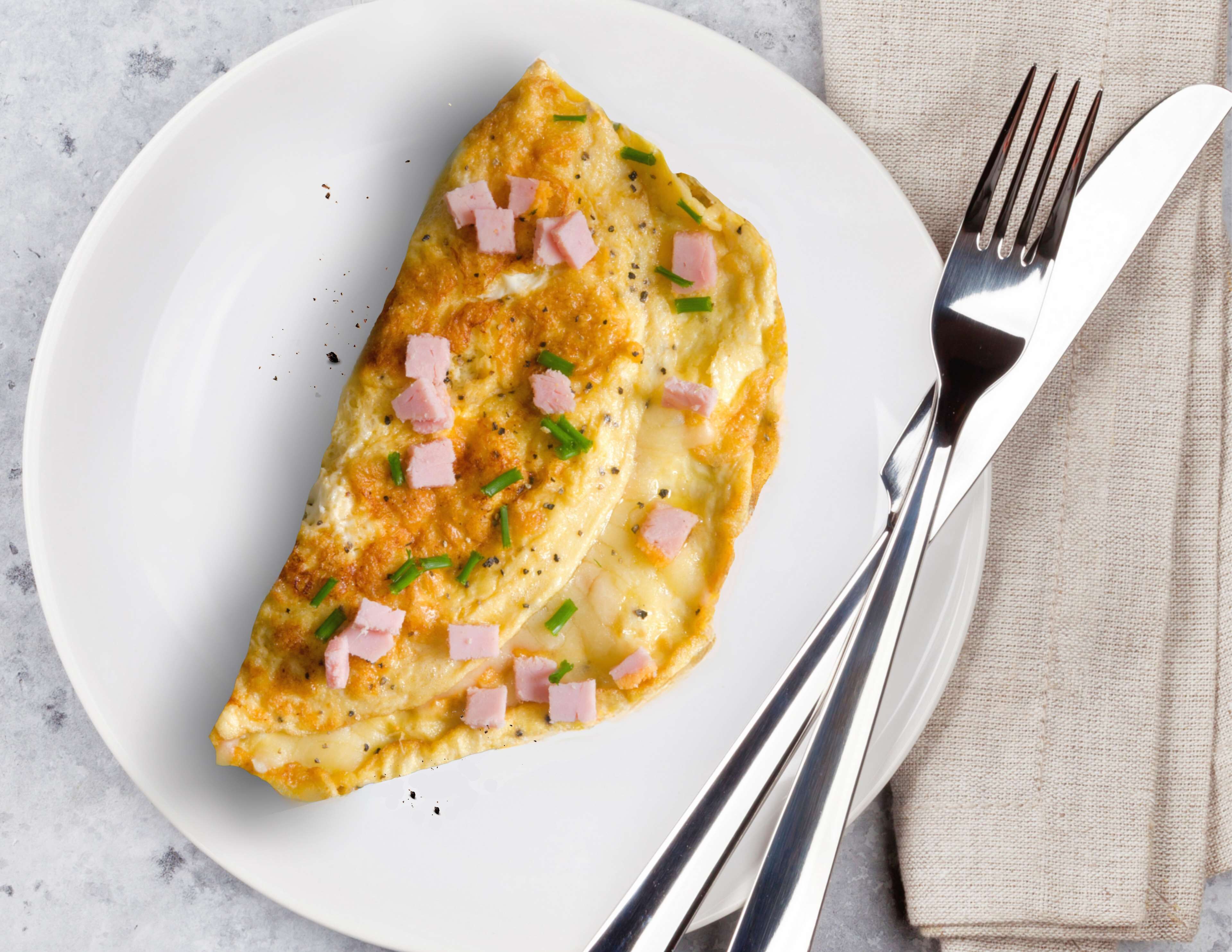 A plate with a folded omelette garnished with ham and chives, accompanied by a fork and knife on a beige napkin.
