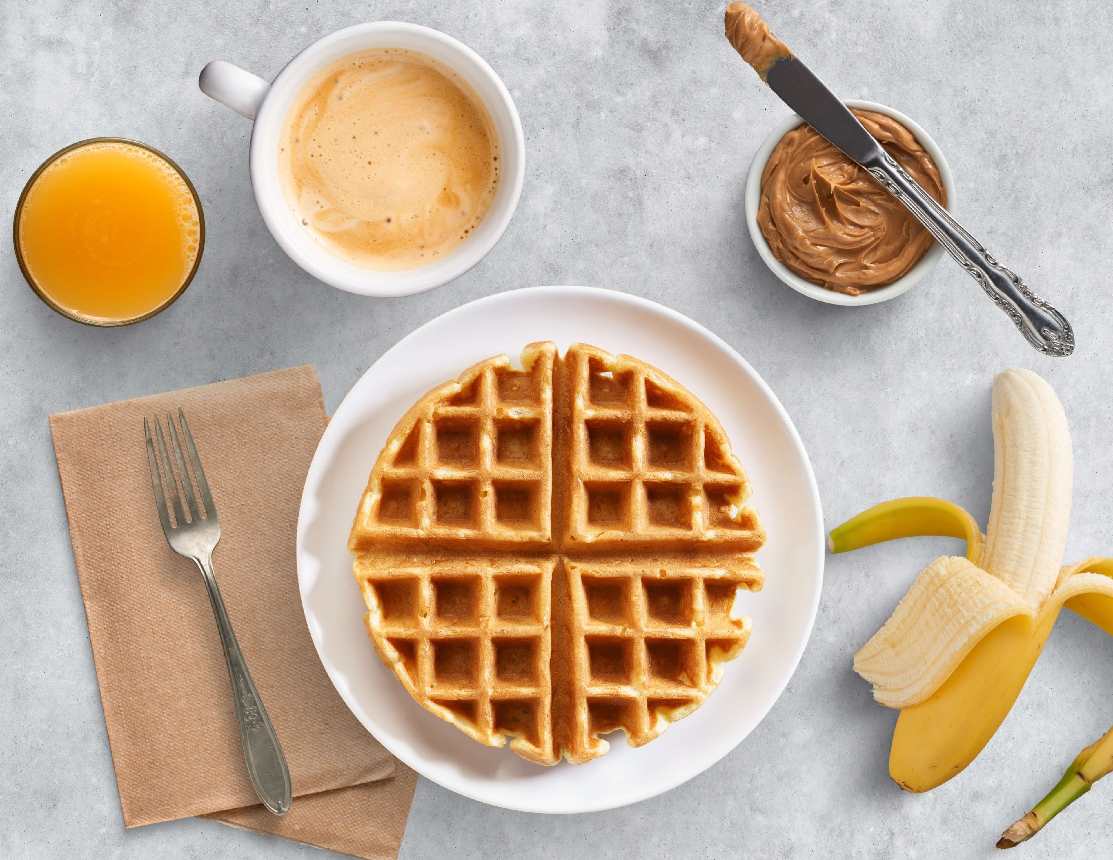 A plate of waffles, a banana, a glass of orange juice, a bowl of peanut butter, and a cup of coffee on a table.