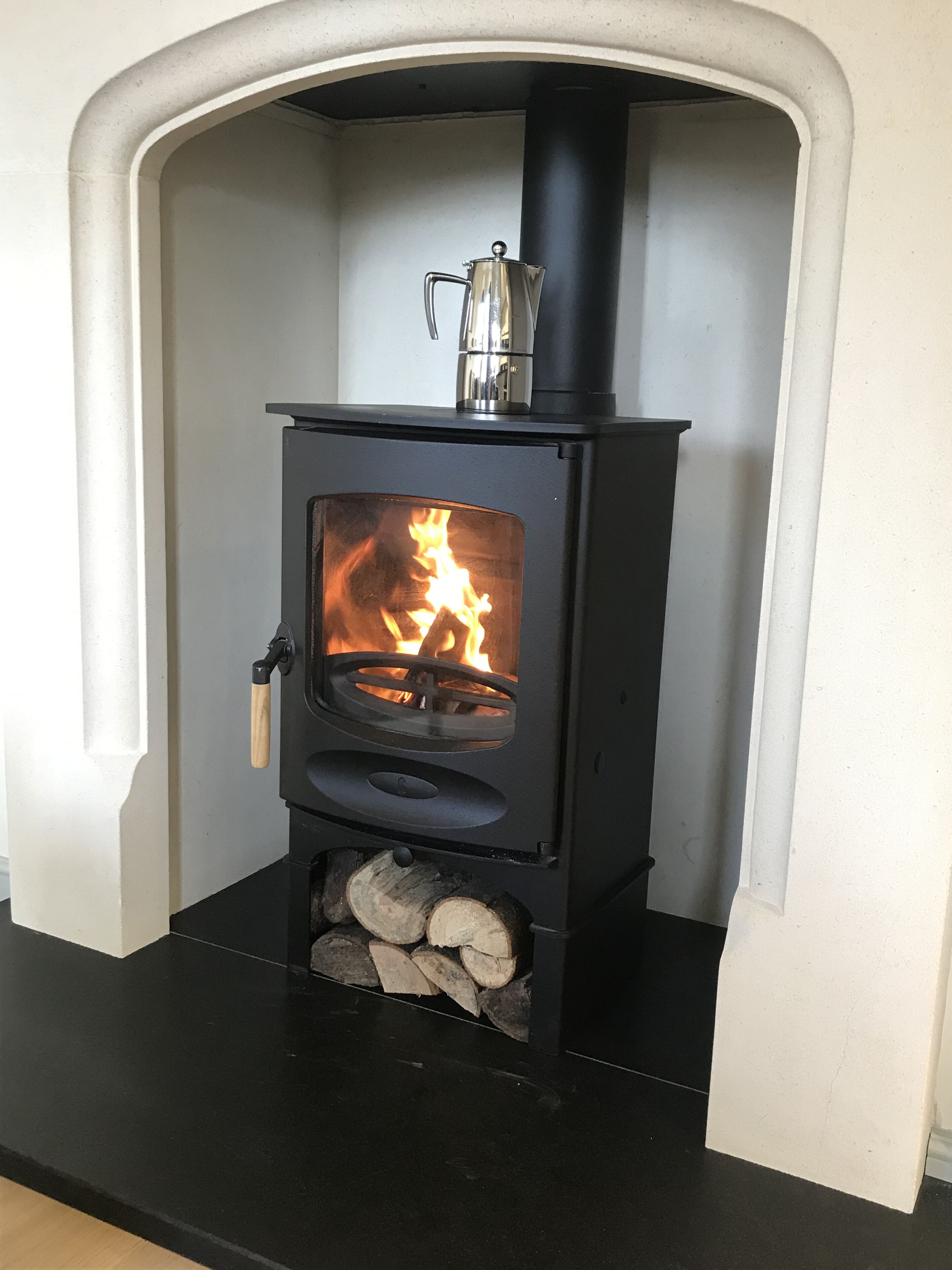 A wood-burning stove with a visible fire, a metal pot on top, and logs stored below.