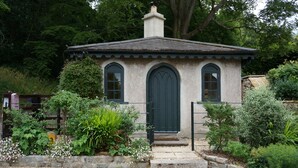 A small, rustic building with a green door and windows, surrounded by lush greenery and a gravel path.