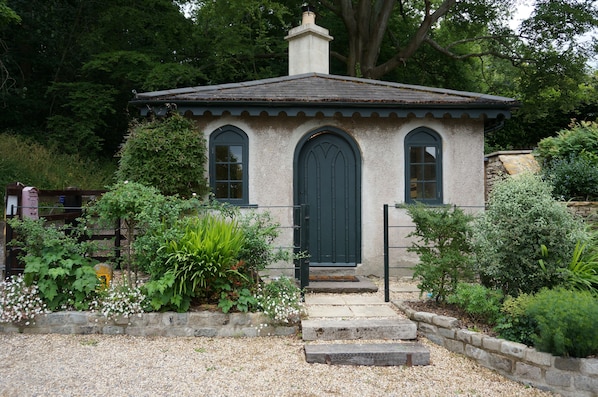 A small, rustic building with a green door and windows, surrounded by lush greenery and a gravel path.