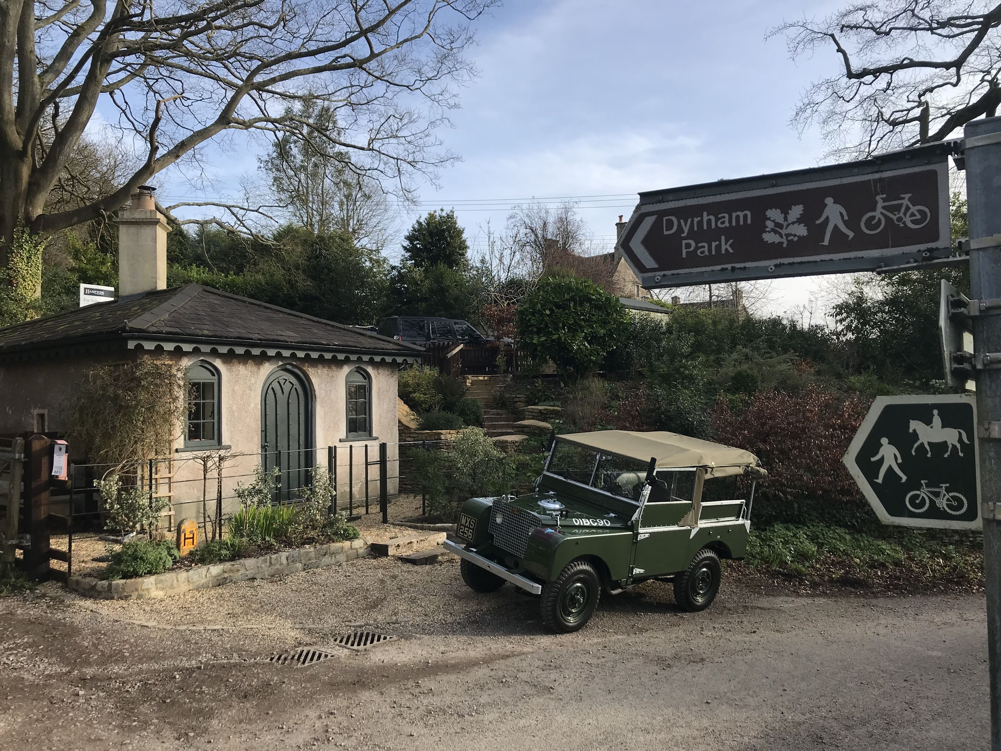 Un edificio de estilo arquitectónico tradicional, un vehículo verde antiguo estacionado al frente, y un cartel con indicaciones hacia Dyrham Park.