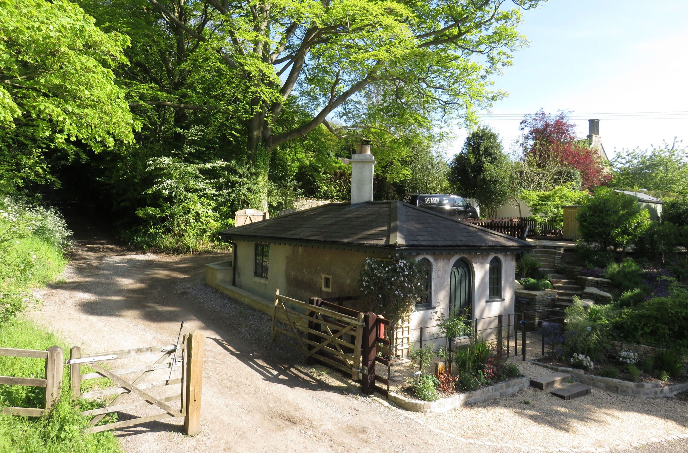 A rustic house with a stone pathway leading to a gated entrance, surrounded by lush greenery and a small garden.