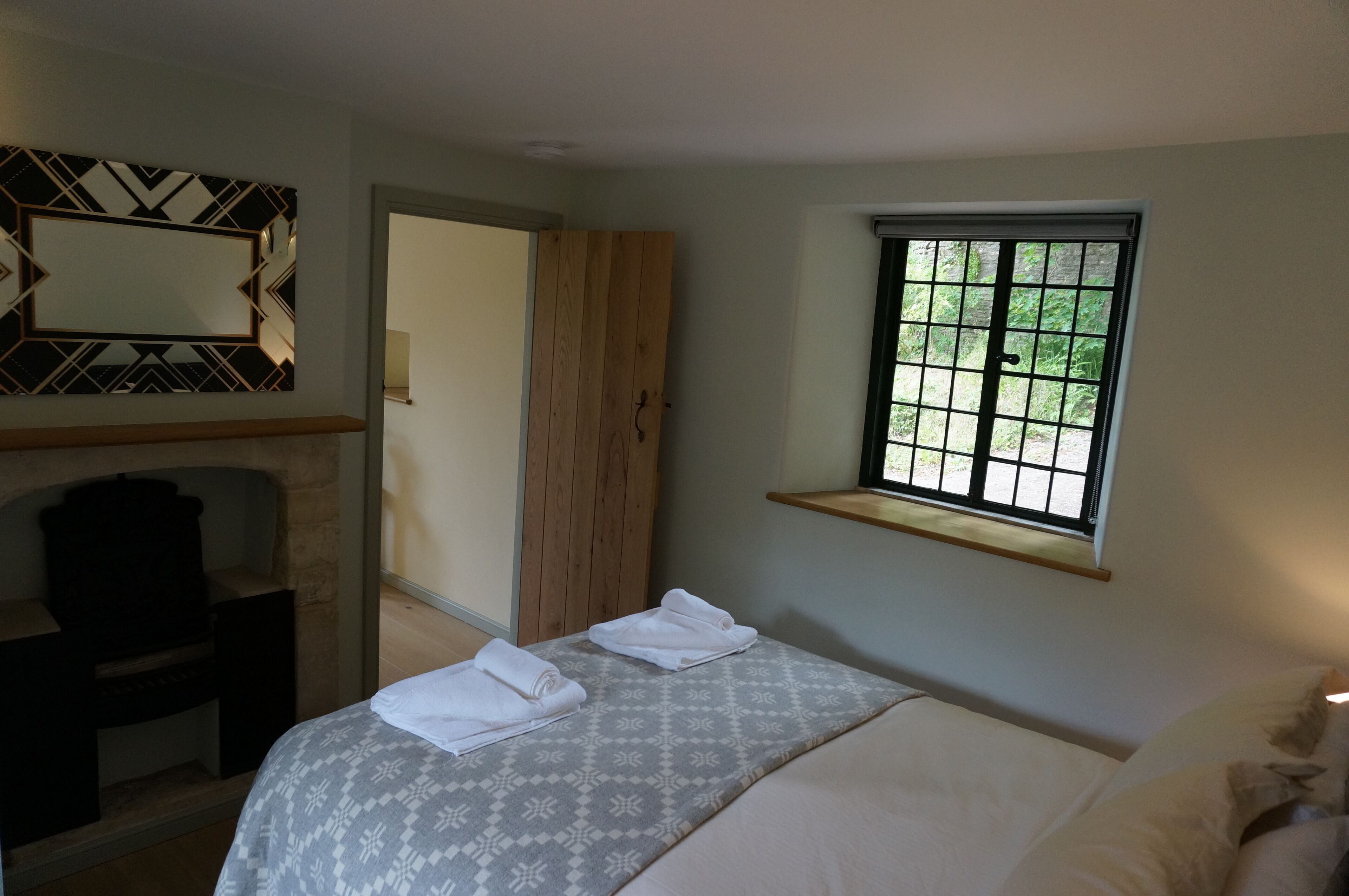 A bedroom with a bed, a wooden wardrobe, a mirror, and a window with a view of greenery.