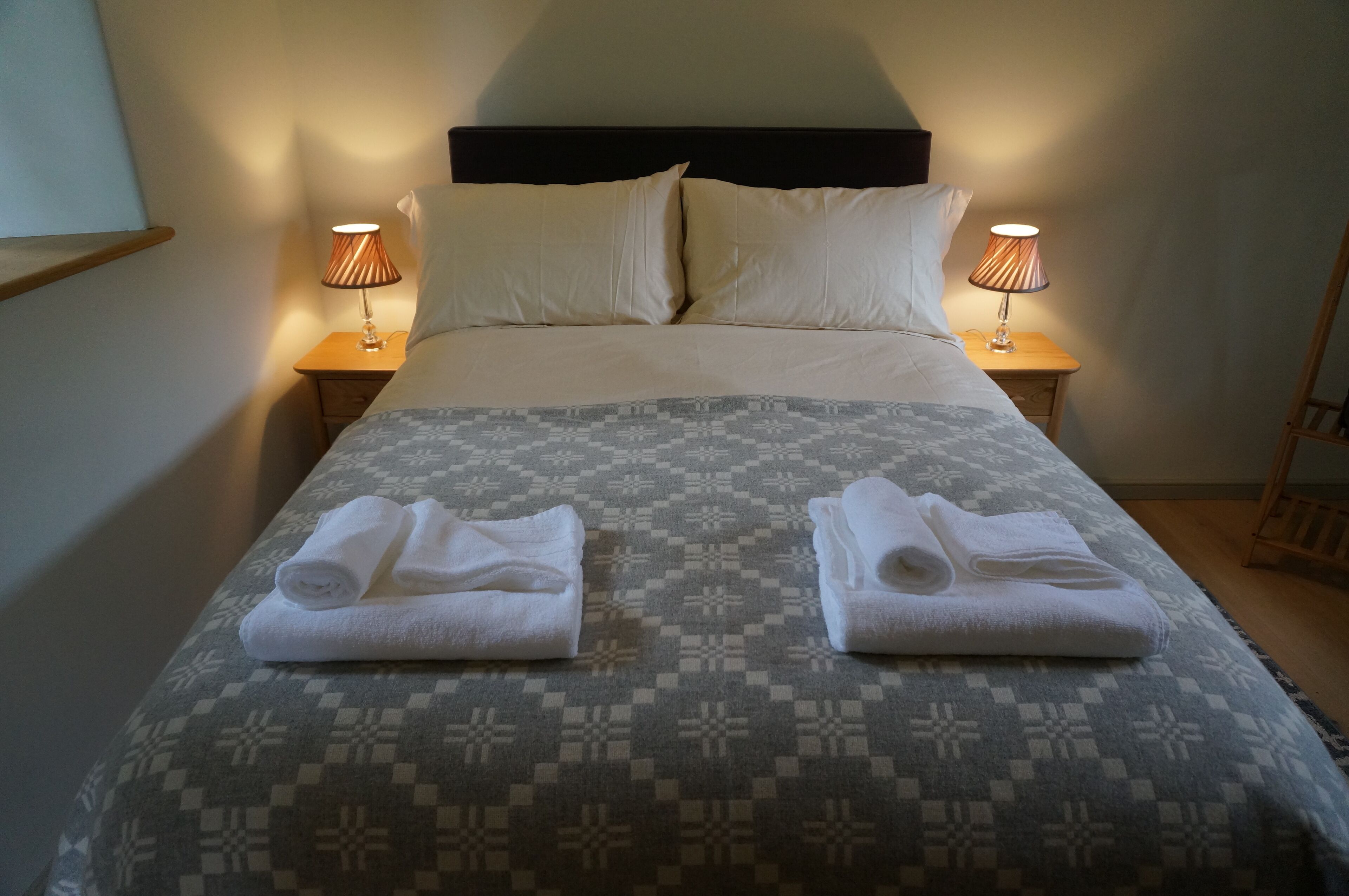 Uma cama bem arrumada com um edredom estampado, dois toalhas enroladas sobre ela e duas luminárias de cabeceira em mesas de madeira.