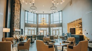 A spacious lobby with high ceilings, large windows, and a central chandelier. There are multiple seating areas with sofas and armchairs. - voco Saranac Lake NY - Waterfront by IHG (Saranac Lake)