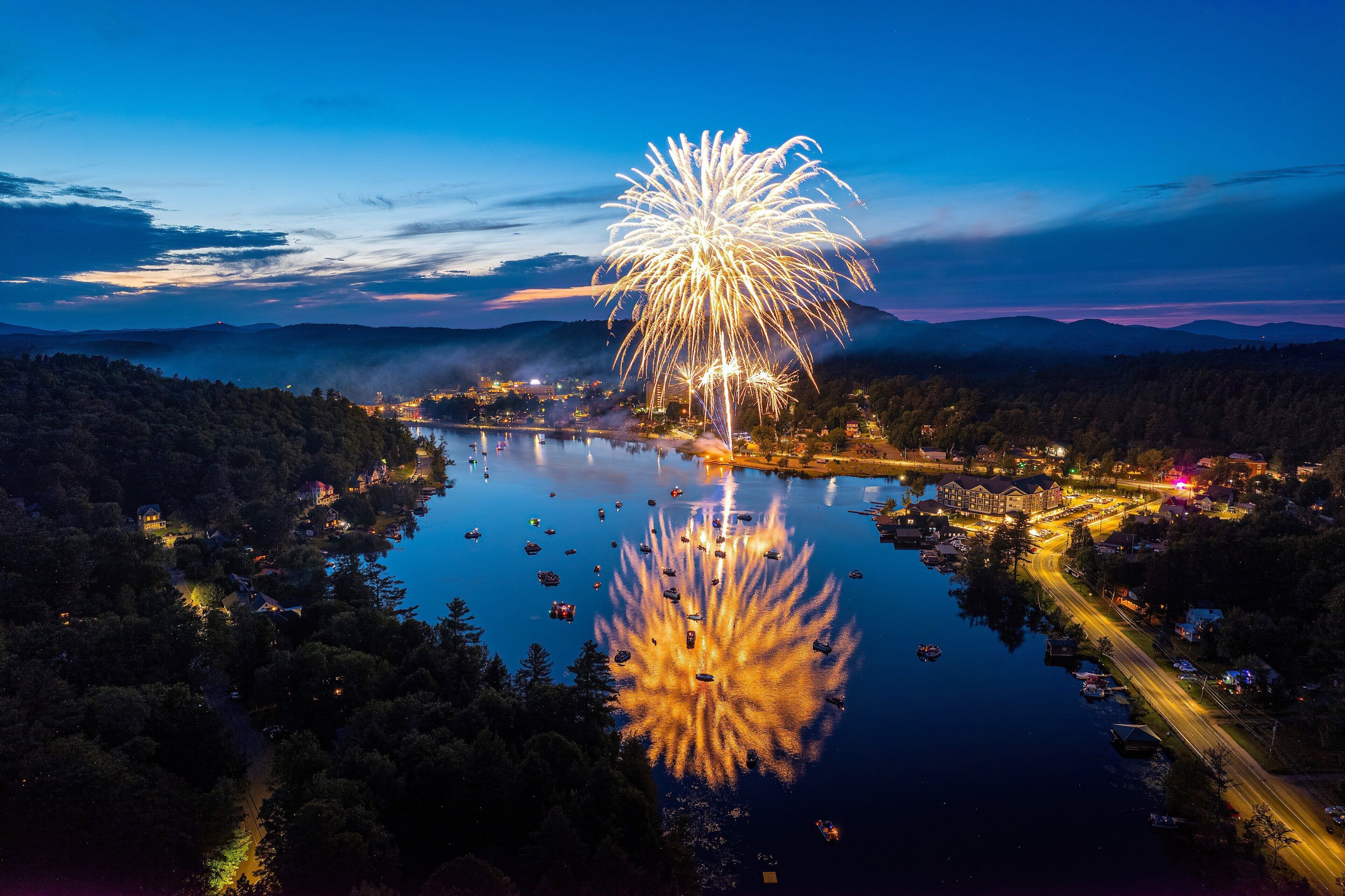 Un pueblo junto al lago de noche, con fuegos artificiales reflejándose en el agua.