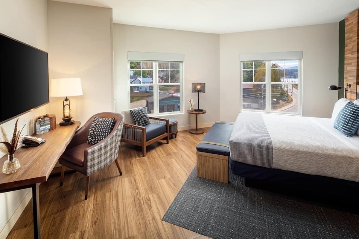 A hotel room with a large bed, a desk with a TV, a chair, two windows offering a view, and a small table with a lamp.