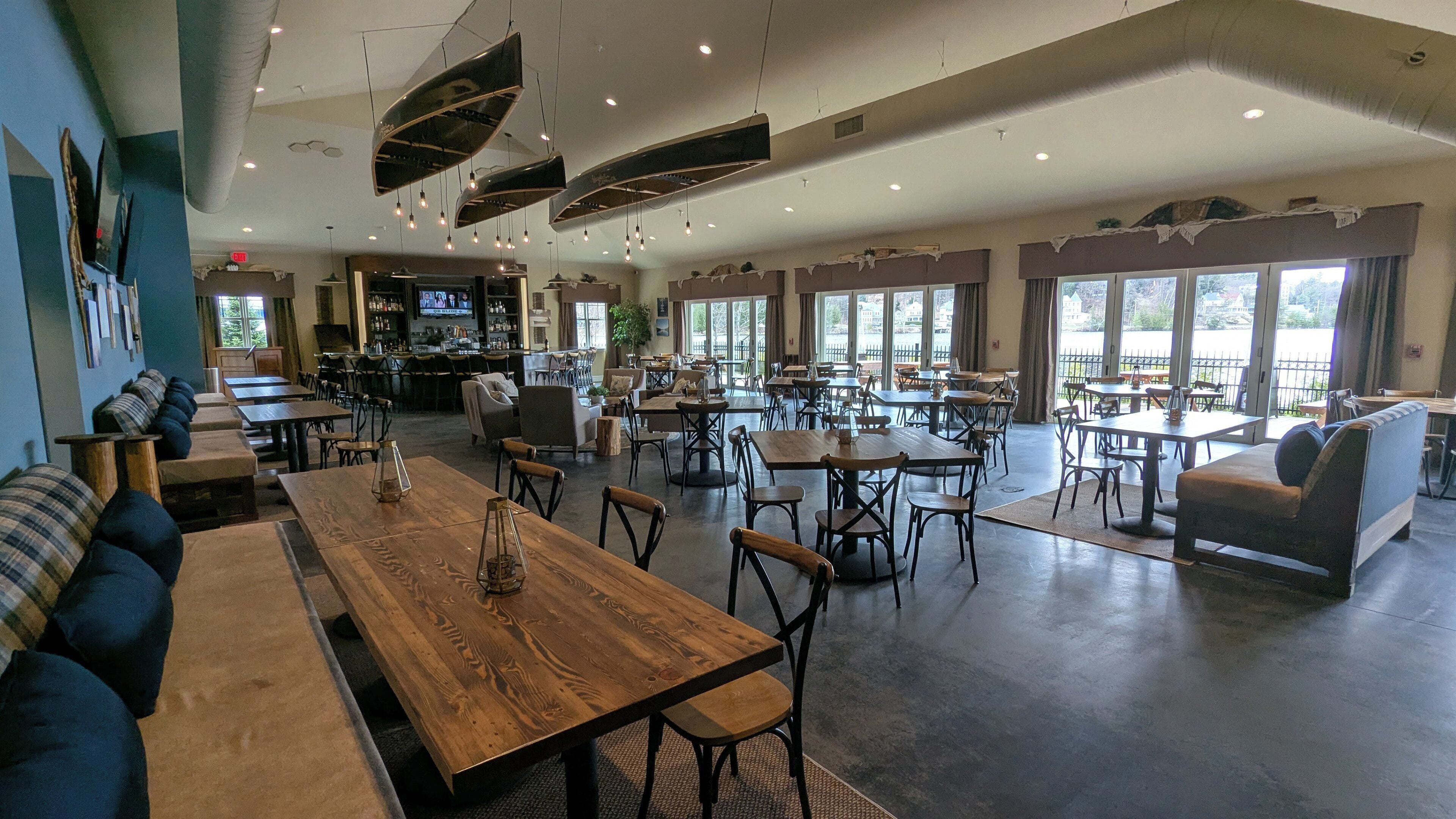 Un amplio comedor interior con varias mesas y sillas, una zona de bar con televisor, y grandes ventanales que ofrecen luz natural.