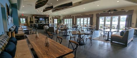 A spacious indoor dining area with multiple tables and chairs, a bar area with a television, and large windows providing natural light.