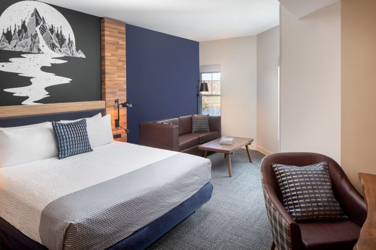 A modern hotel room with a large bed, a brown leather sofa, a small coffee table, and a wall-mounted TV.