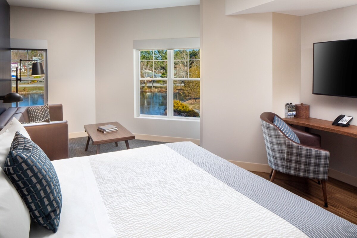 A hotel room with a large bed, a desk, a chair, a TV, and a window with a view of a body of water.