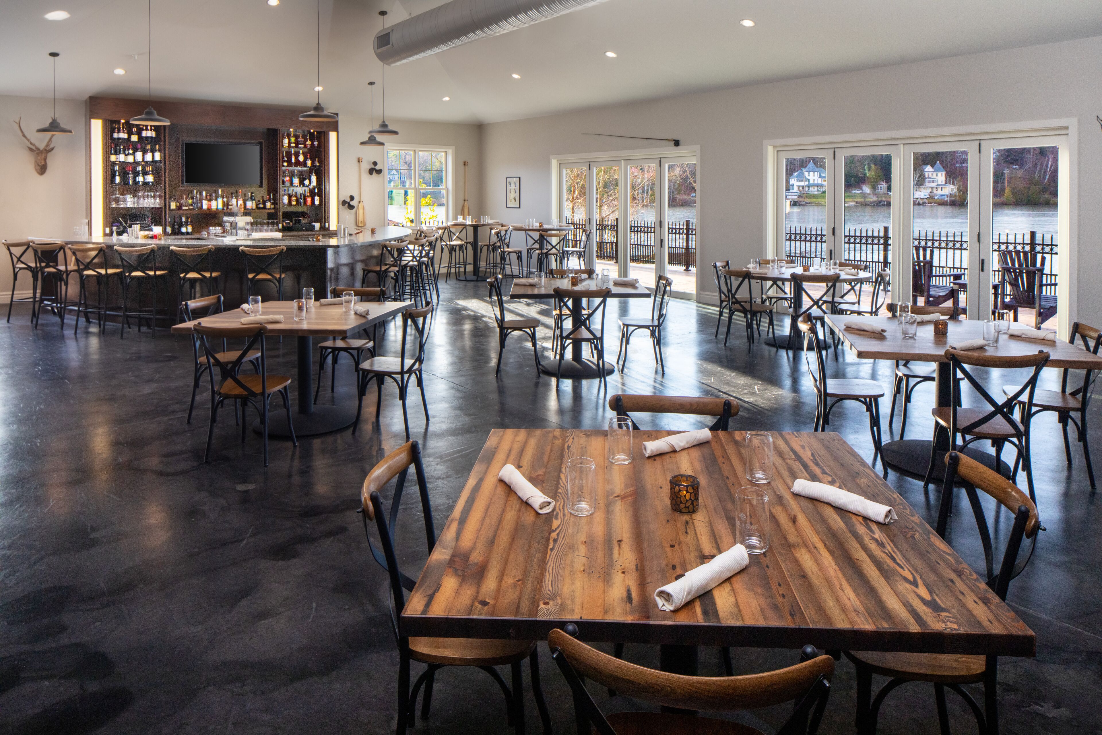 A spacious restaurant with multiple wooden tables and chairs, a bar area, and large windows offering views of a lake.