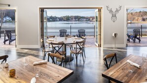 A dining area with wooden tables and chairs, a view of a lake, and a deer head decoration on the wall. - voco Saranac Lake NY - Waterfront by IHG (Saranac Lake)