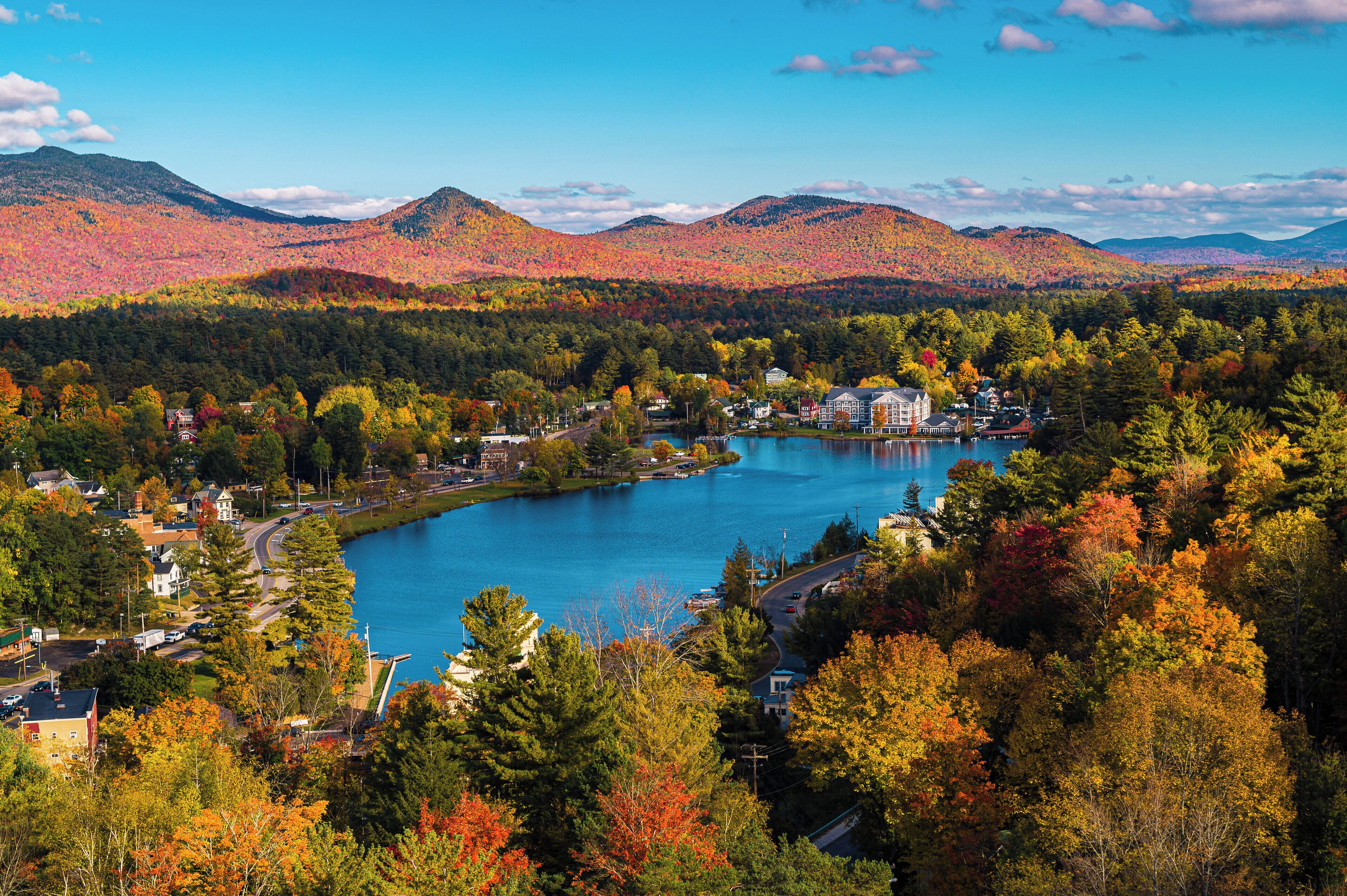 A lakeside town with houses and a large building, surrounded by autumn-colored trees and mountains.