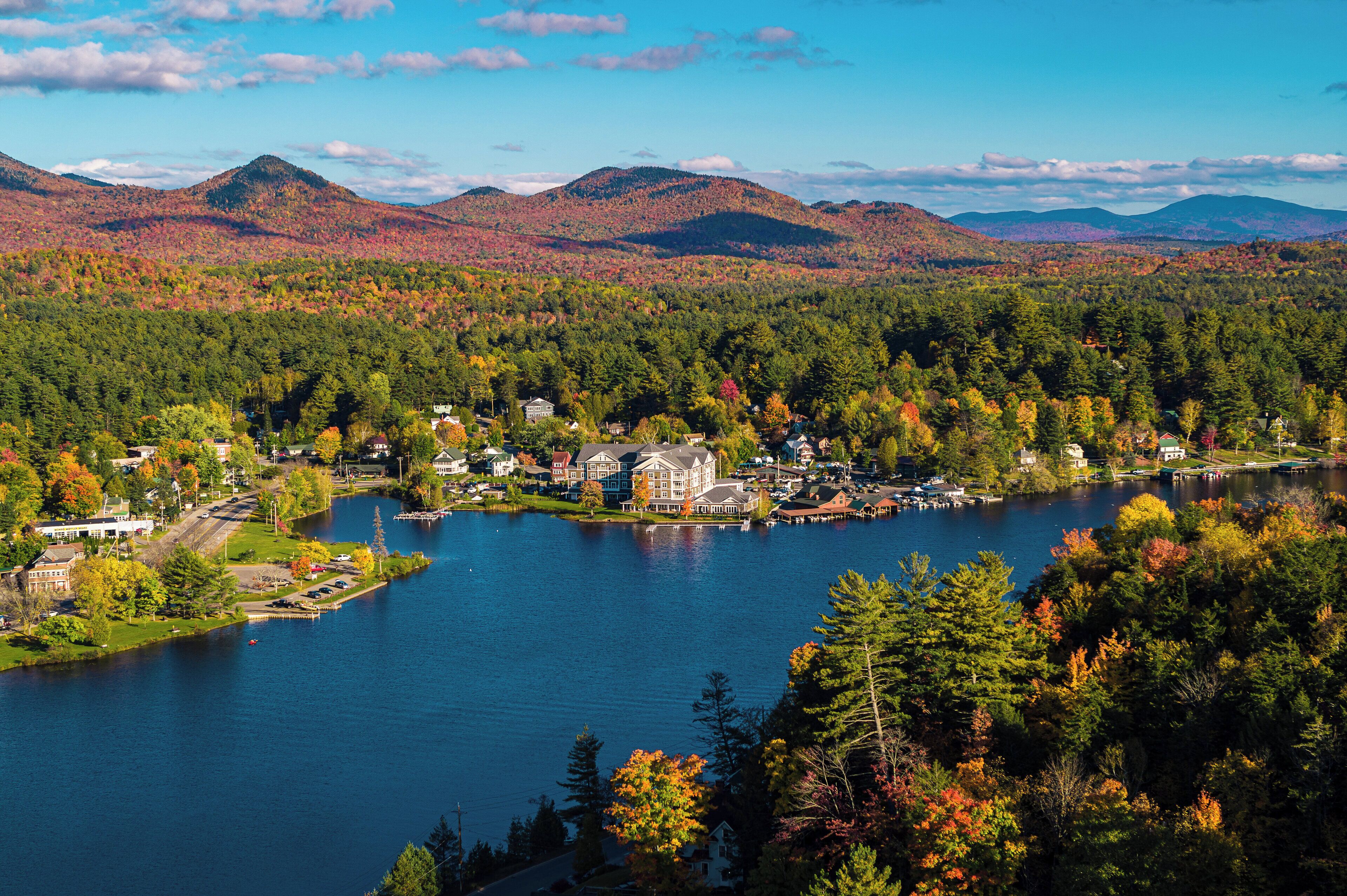 A lakeside town with houses and a large building, surrounded by mountains and autumn foliage.