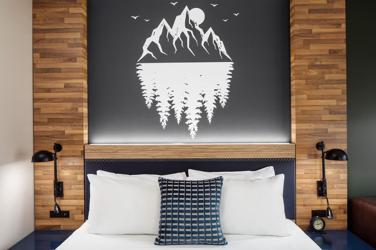 A hotel room with a wooden headboard, a bed with white and patterned pillows, a wall-mounted artwork of mountains and trees, and two bedside lamps.