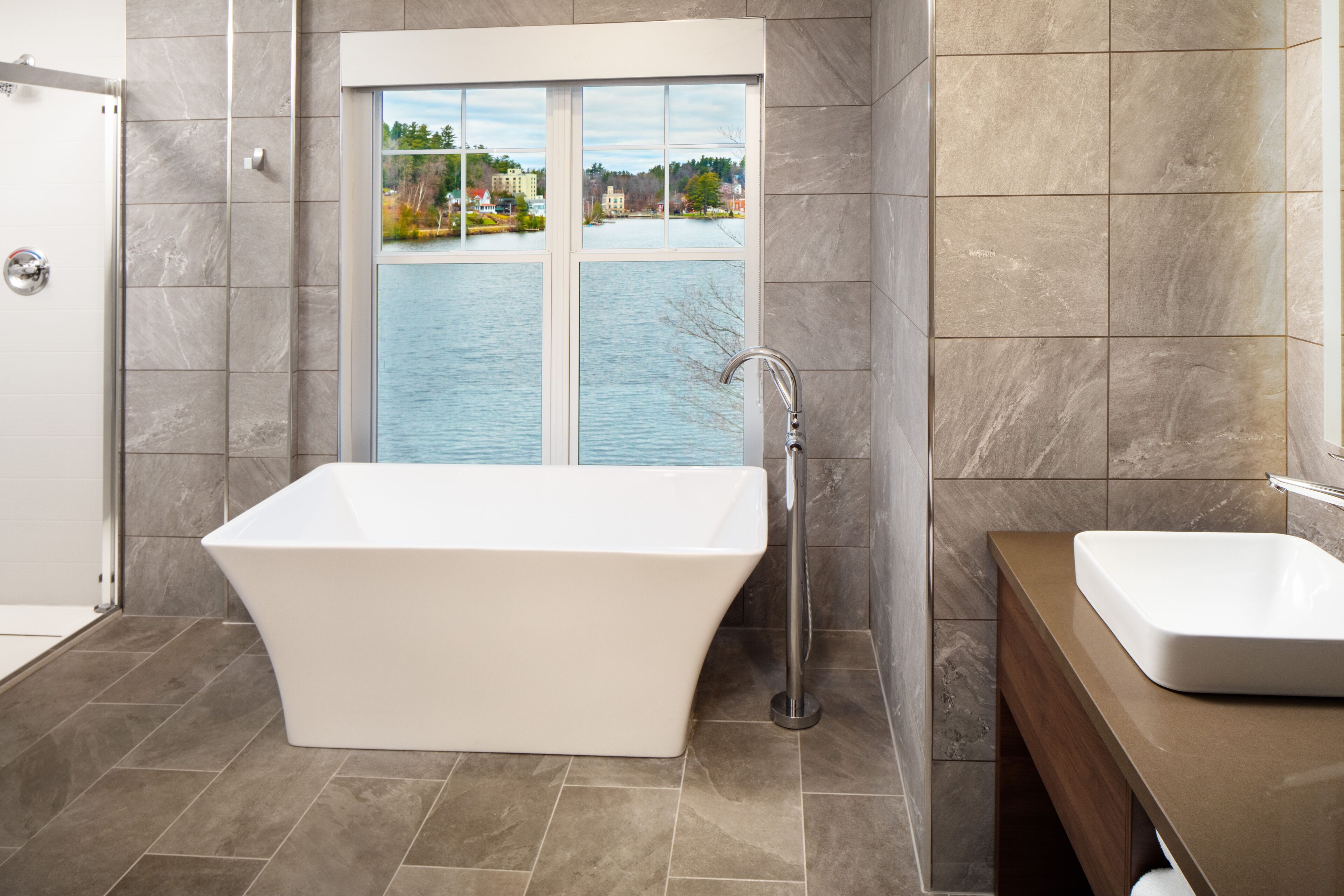 A modern bathroom with a freestanding bathtub, a window with a view of a lake and buildings, and a wooden vanity with a sink.