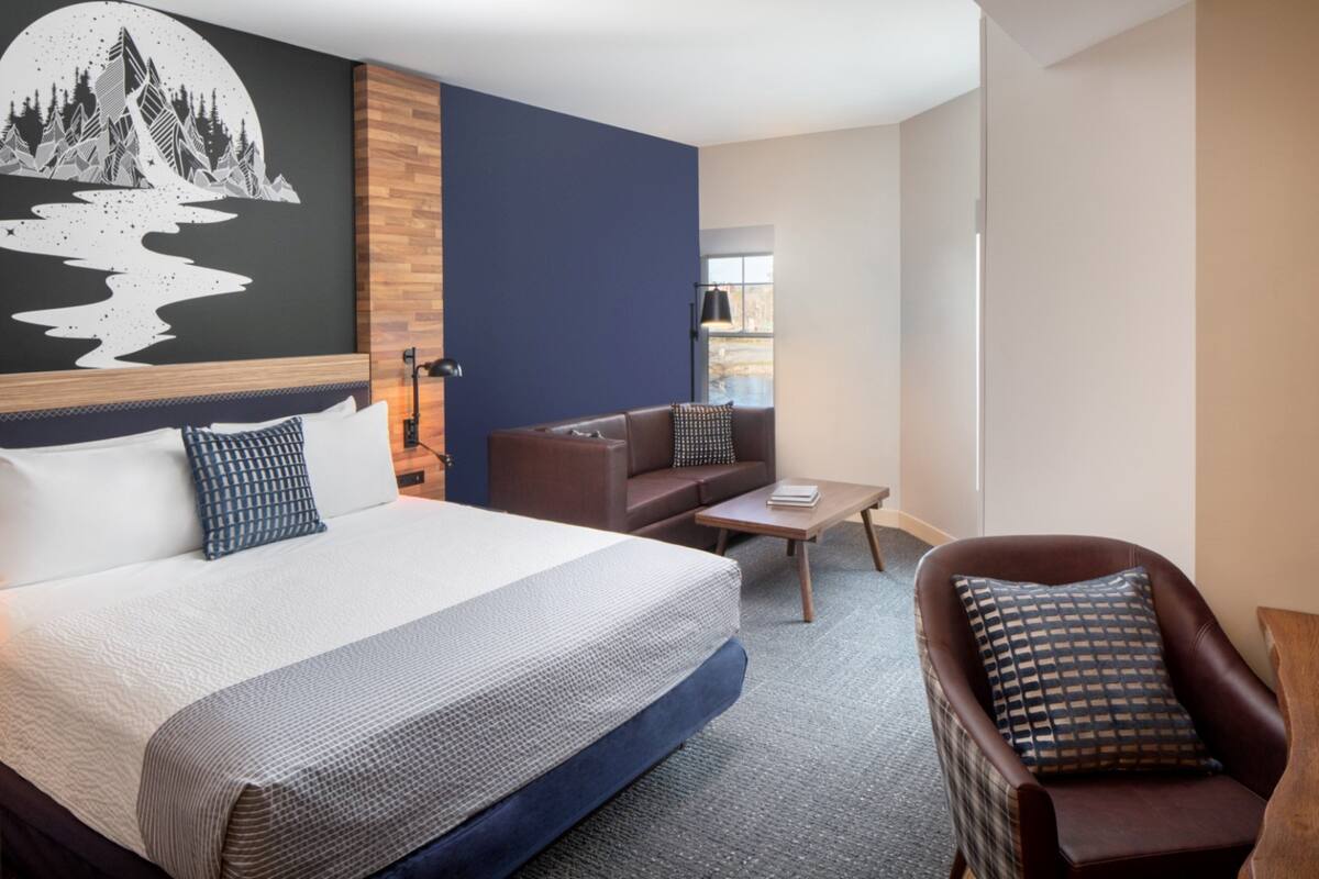 A modern hotel room with a large bed, a brown leather sofa, a small coffee table, and a wall-mounted TV.