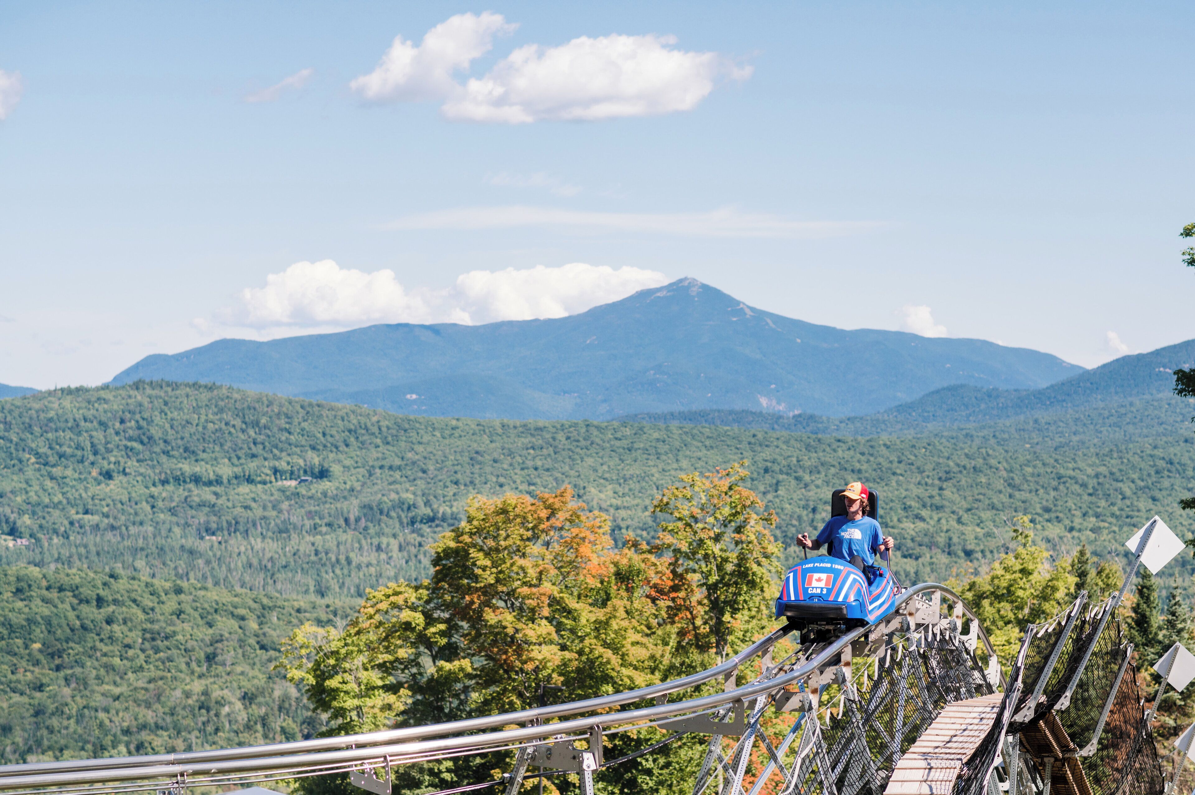 A chairlift with a person seated, overlooking a mountainous landscape with dense forests and a prominent peak.