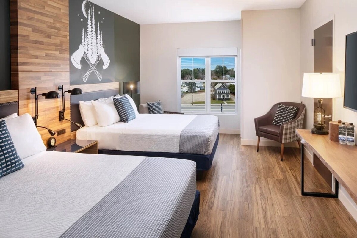 A hotel room with two beds, a desk with a lamp, a chair, and a large window offering a view of the outside.