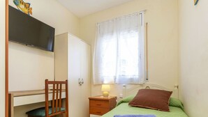Un dormitorio con cama, escritorio, silla, armario, ventana con cortinas y televisión en la pared.