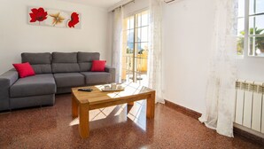 Een woonkamer met een grijze bank, rode kussens, een houten salontafel en een groot raam met gordijnen.