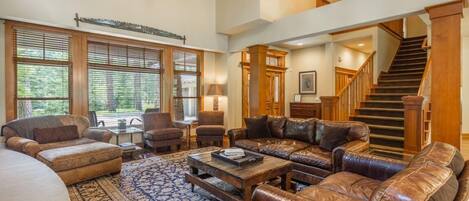 A spacious living room with a large sectional sofa, a coffee table, and a patterned rug. There is a staircase leading to an upper level, and large windows with blinds.