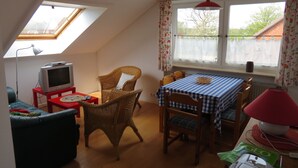 A cozy room with a sloped ceiling, a television on a red stand, a wicker chair, a dining table with a checkered tablecloth, and a window with floral curtains.