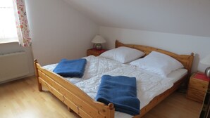 A wooden bed with white bedding and blue towels, a nightstand with a lamp, and a window with floral curtains.
