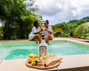 A poolside setting with a wooden table holding a bottle, glasses, and fruit.