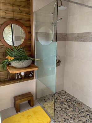 A modern bathroom with a glass-enclosed shower, a wooden bench, a round mirror, and a decorative stone basin.