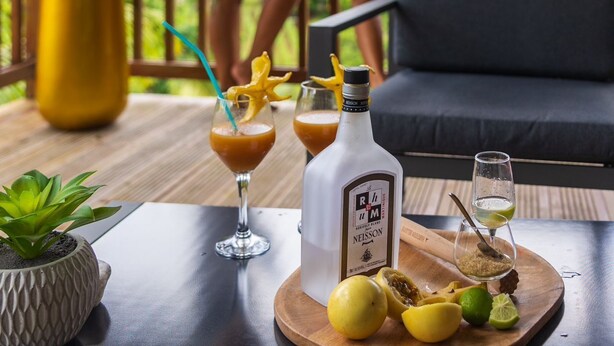Eine Holzdielen-Terrasse mit einer Couch und zwei Personen in weißen Roben. Auf dem Tisch stehen Getränke und eine Flasche.