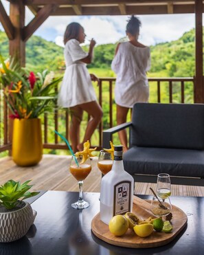 A wooden deck with a sofa and two people in white robes. There are drinks and a bottle on the table. - Les Bungalows de la Sucrerie: spacious, quiet, Bac à Punch, near the southern beaches (Le François)