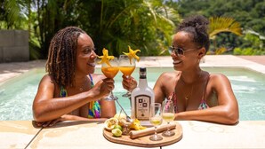 Zwei Personen stoßen mit Getränken an einem Tisch am Pool an, mit einer Flasche Nesson und tropischen Früchten.