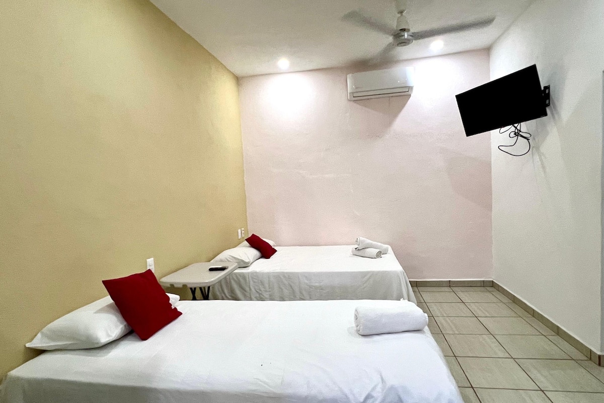 A hotel room with two single beds, a ceiling fan, an air conditioning unit, and a flat-screen TV mounted on the wall.