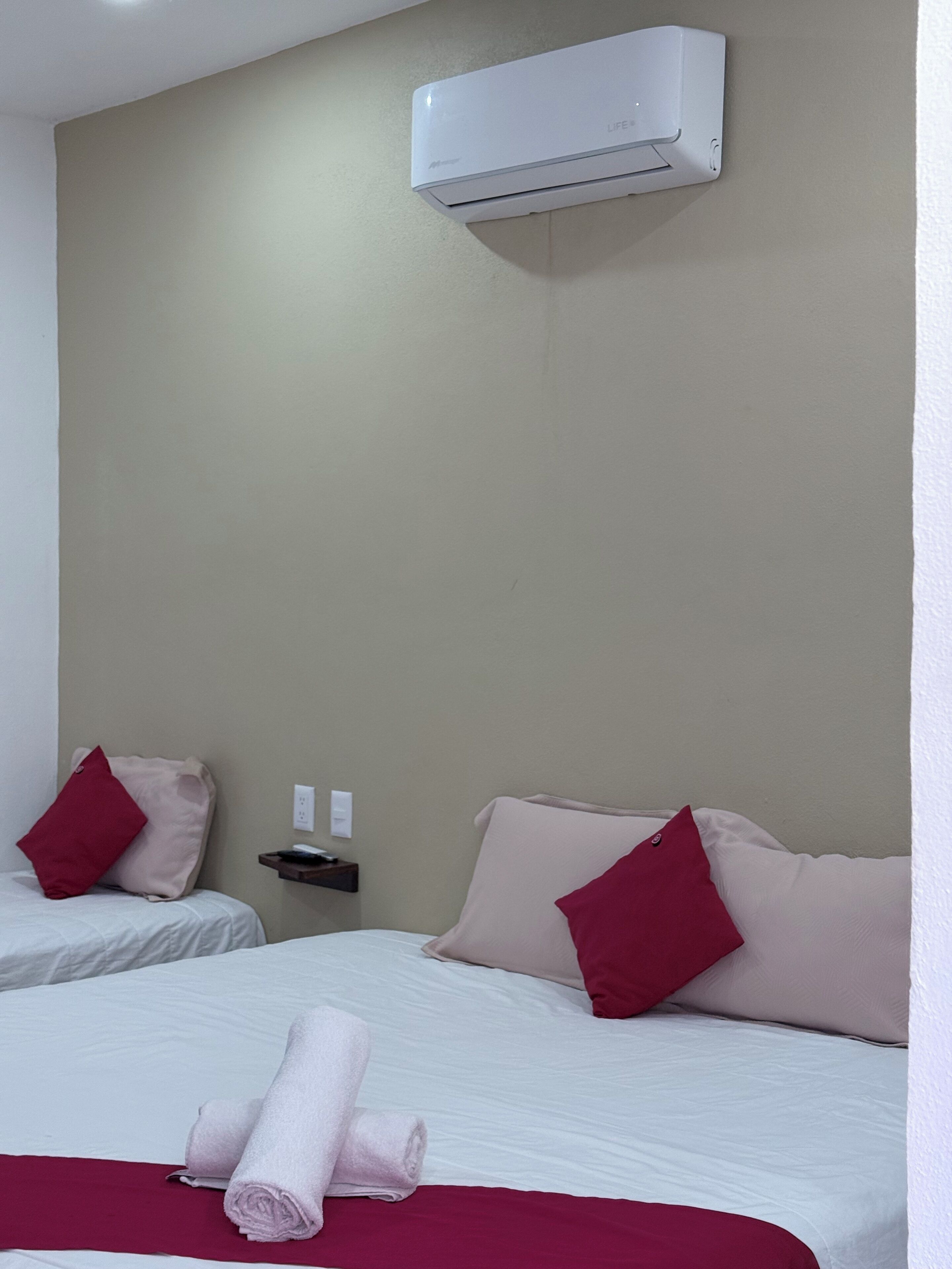 A hotel room with two beds, each with a red and beige pillow, a white bedspread, and a rolled towel on the bed. There is a small table with a phone and a power outlet on the wall.
